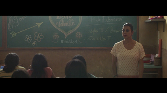A woman in a white patterned shirt stands before a green chalkboard with Valentine's Day decorations.