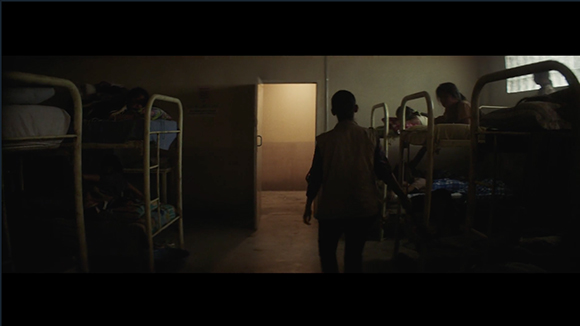 A person walks through a dimly lit dormitory lined with bunk beds toward an open doorway.