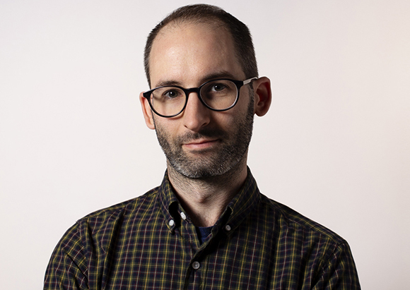 A man with short dark hair, glasses, and a beard wearing a plaid shirt against a white background.