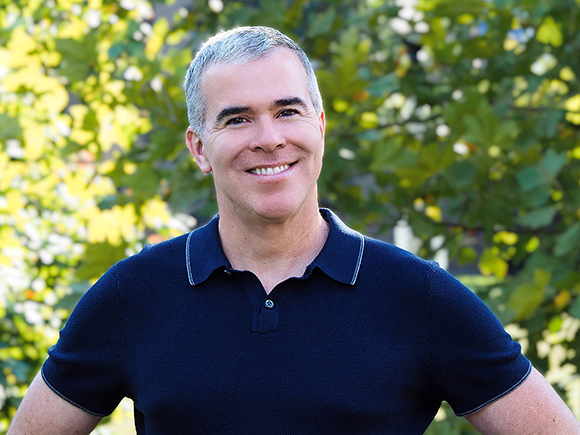 A smiling man with short gray hair wearing a black polo shirt against a leafy green background.