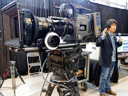 A large professional cinema camera with a long lens mounted on a heavy-duty tripod.