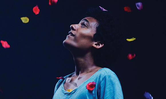 A person with dark curly hair looks upward against a dark background as colorful flower petals fall.