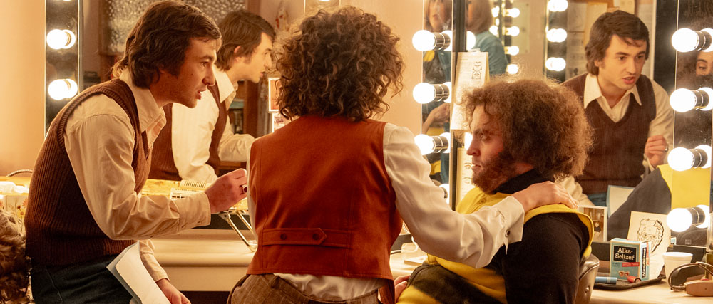 A man in a brown vest applies makeup to a man with curly hair in a dressing room mirror.