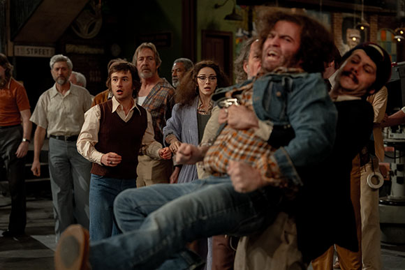 A crowd of people with surprised expressions watches a man being carried through a room.