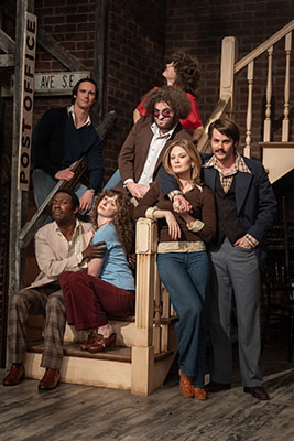 Seven actors in vintage clothing pose on a wooden staircase against a brick wall.