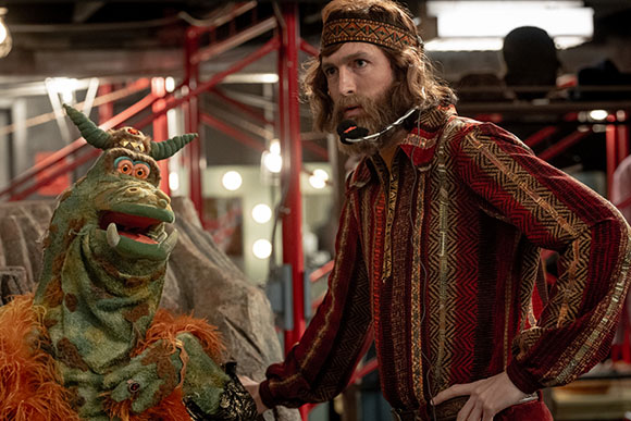 A man with a beard and headband stands beside a large green and orange puppet in a studio setting.