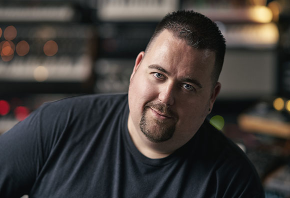 A man with short dark hair and a goatee wearing a black t-shirt, looking toward the camera.