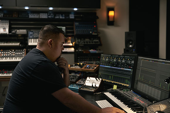 A man in a black shirt sits at a desk in a music studio filled with synthesizers and audio equipment.