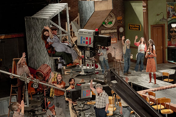 A theatrical stage set showing actors and crew members operating cameras and equipment.