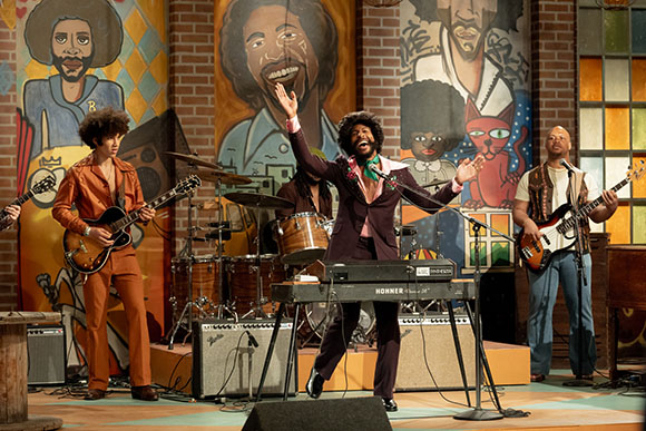 Four musicians perform on a stage decorated with large, colorful character murals.