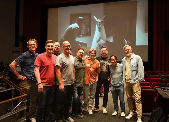 Nine men standing in a row in a dark movie theater with a large screen in the background.