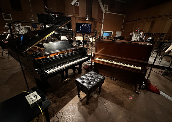 A black grand piano and a brown upright piano sit in a dimly lit music studio with various equipment.
