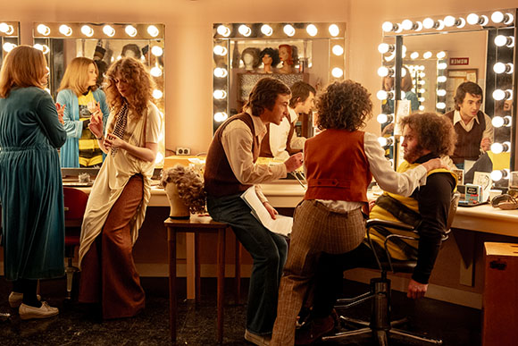 Several people in vintage clothing prepare in a dressing room filled with lit vanity mirrors.