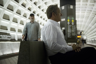 A man in a white shirt sits on a concrete ledge in a subway station while another man walks in the background.