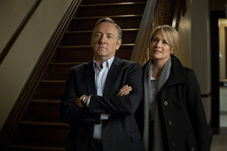 A man in a dark suit with arms crossed stands next to a woman in a black coat and grey scarf.