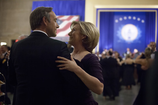 A man in a dark suit and a woman in a purple dress dance closely in a ballroom.