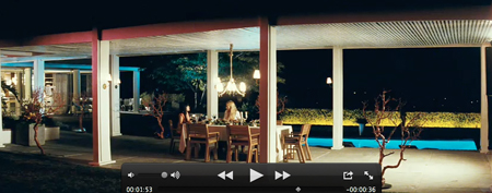 People dining at a long table under a covered patio at night near a glowing blue pool.