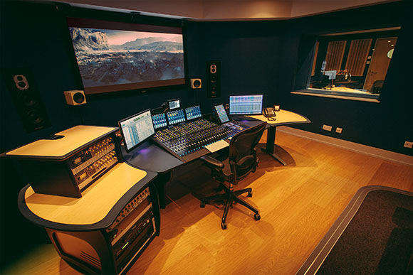 A professional recording studio featuring a large mixing desk, computer monitors, and a large video screen.