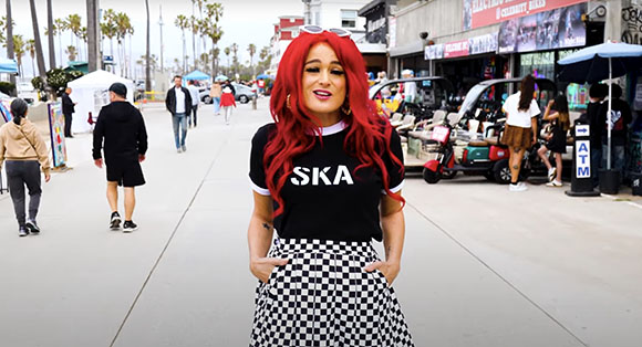 A woman with long red hair in a black t-shirt and checkered skirt stands in a busy outdoor plaza.