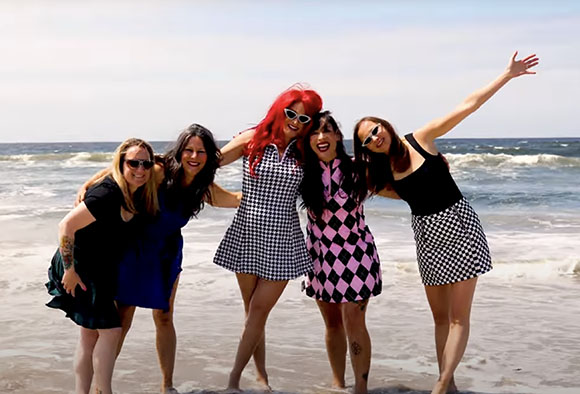 Five women in patterned dresses posing and smiling on a sunny beach with ocean waves.