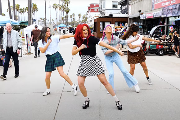Four women in casual clothing dance together on a sunny outdoor walkway.