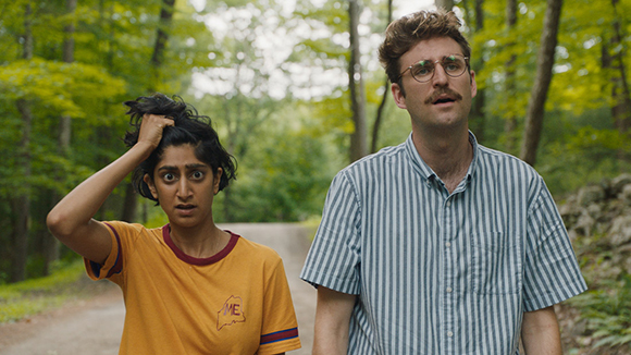 A woman in a yellow shirt and a man in a striped shirt look surprised on a path through a forest.