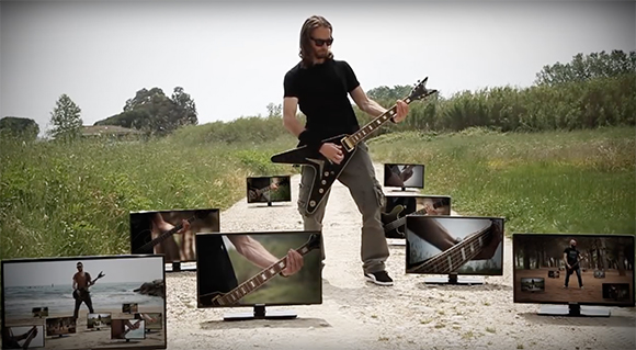 A man in black playing a black electric guitar on a dirt path surrounded by multiple TV screens.