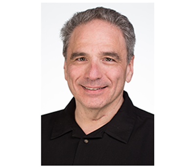 A middle-aged man with graying hair and a black polo shirt smiling against a white background.