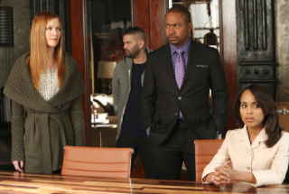 A woman in a white blazer sits at a table while three others stand behind her in an office setting.