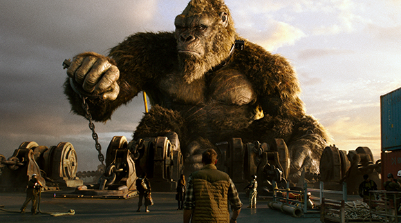 A giant gorilla stands over several small people on a dock at sunset.