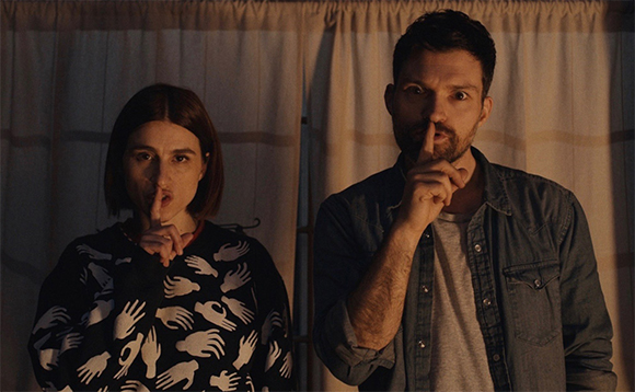 A man and a woman stand in front of curtains, both holding a finger to their lips in a silence gesture.