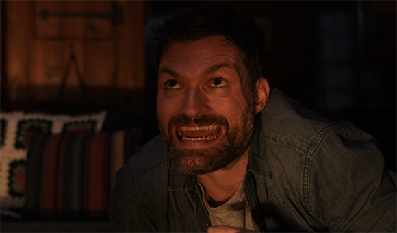 A bearded man looks upward with a tense, grimacing facial expression in dim lighting.