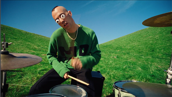 A man wearing googly eyes over his eyes plays a drum kit in a green, hilly field under a blue sky.