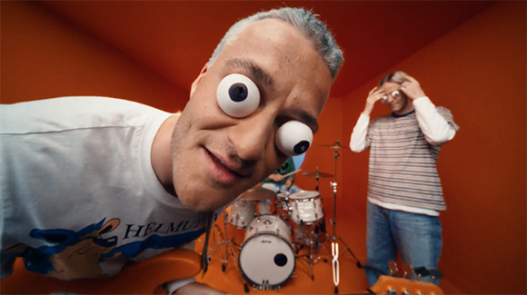 A man with large googly eyes on his face leans toward the camera in an orange room near a drum set.