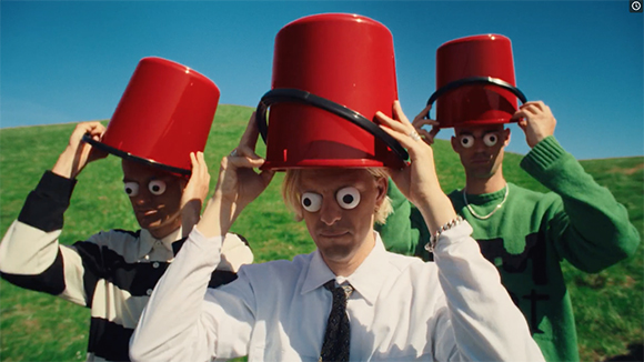 Three men in a field wearing red buckets on their heads with large googly eyes over their eyes.
