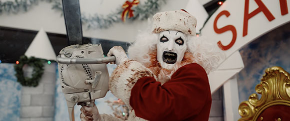 A person in white clown makeup and a red suit holds a large chainsaw.