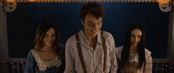 A man in suspenders stands between two women, all three smiling against a dark background with lights.