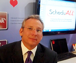 A man wearing glasses and a suit sits in an office in front of a computer screen showing software branding.