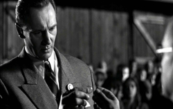 A man in a dark suit and tie looks downward with a serious expression in a black and white film still.