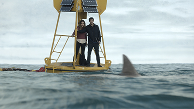A man and woman stand on a yellow buoy in the ocean near a shark fin.