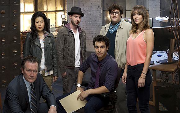 Five people pose together in a cluttered office with filing cabinets and desks.