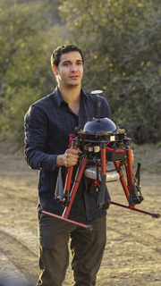A man in a dark button-down shirt carries a large red and black multirotor drone outdoors.