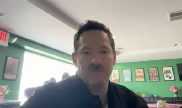 A man with dark hair and a mustache looks directly into the camera in a room with green walls.