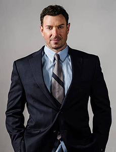 A man wearing a dark navy suit, light blue shirt, and striped tie poses against a plain gray background.