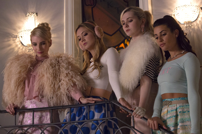Four young women in elegant, feathered, and fur-trimmed outfits standing behind a metal railing.