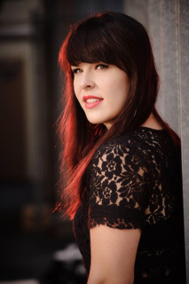 A woman with long dark hair and bangs wearing a black lace top looks over her shoulder.