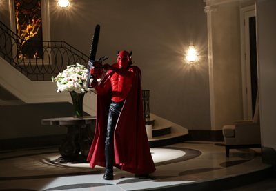 A person in a red devil mask and cape stands in a dimly lit hallway next to a flower arrangement.