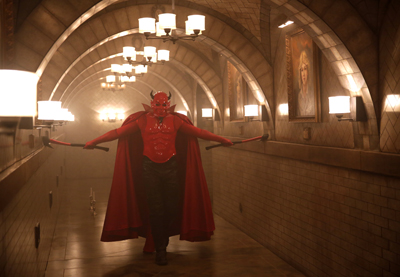 A person in a red devil costume with a cape and horns walks down a dimly lit, arched tiled hallway.