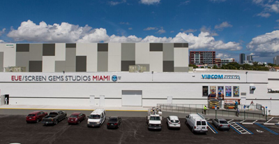 A long white studio building with gray patterned panels under a blue sky with white clouds.