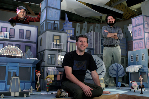 Three men pose behind and among miniature blue and grey city buildings and small character figures.